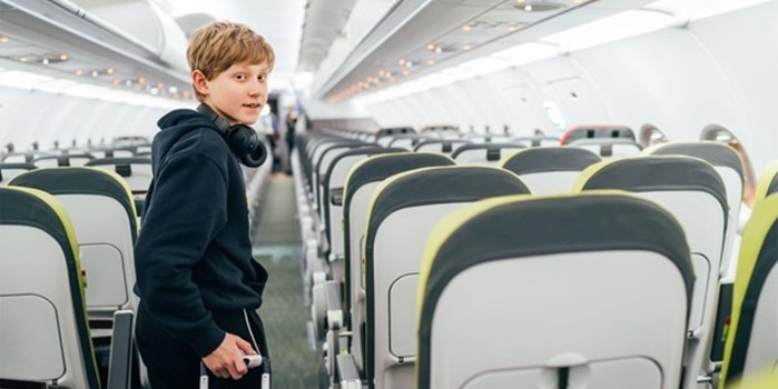 A teenage boy walks down the aisle of a plane pulling a suitcase behind him